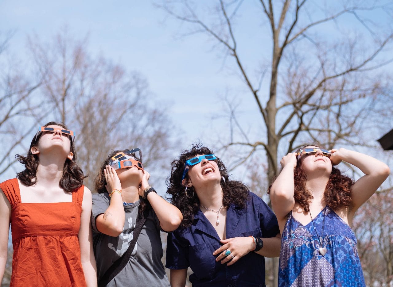 Four women in eclipse glasses looking up under bare branches, April 2024