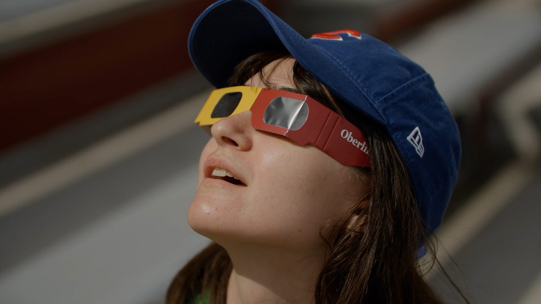 Close-up of a young woman in a baseball cap and red eclipse glasses looking up