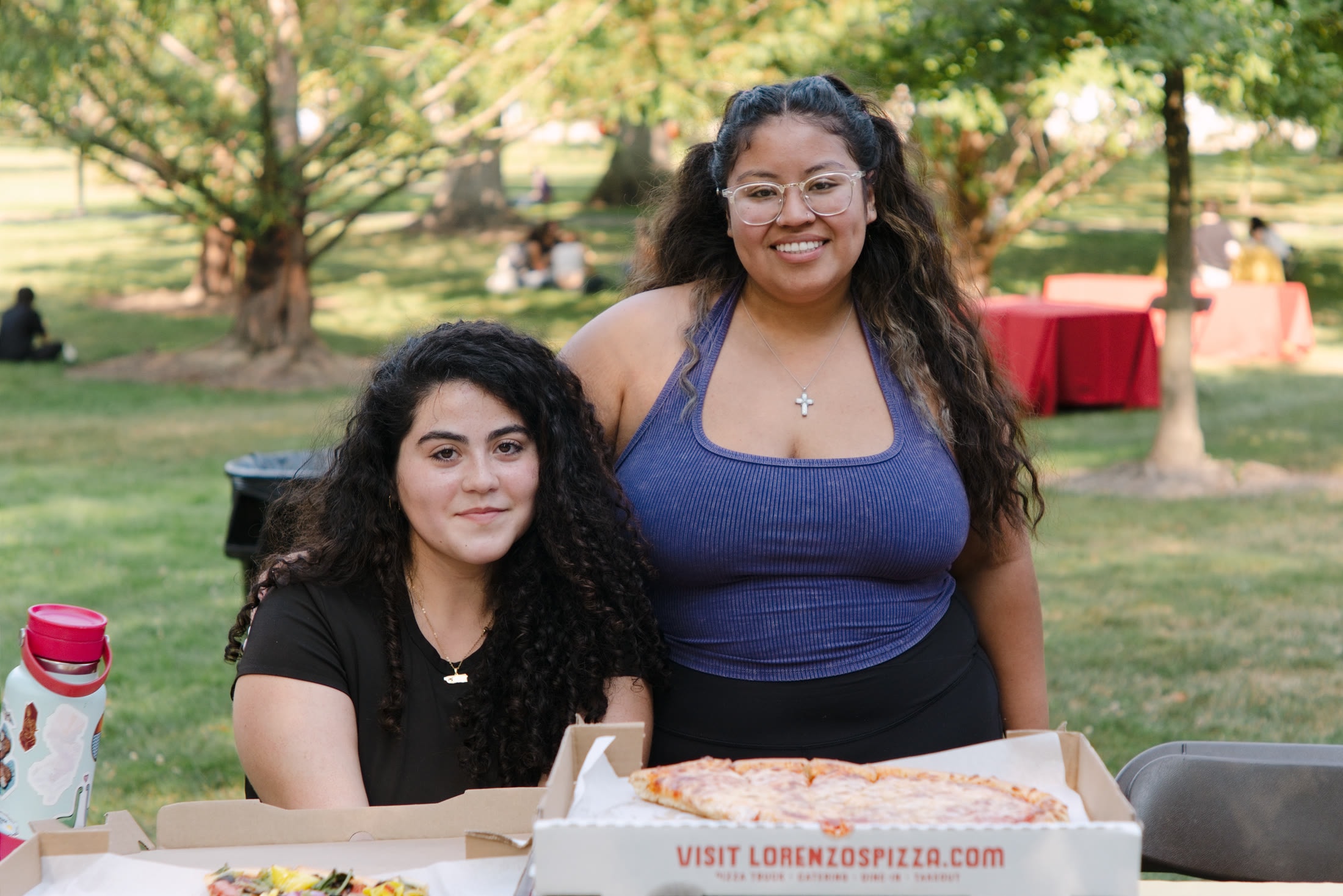 Two students posing behind pizza boxes at an outdoor campus gathering