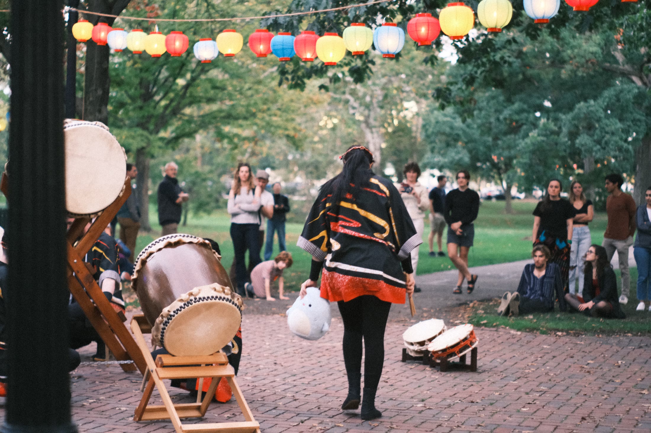 A drummer walks toward instruments beneath colorful lanterns on an Oberlin campus path