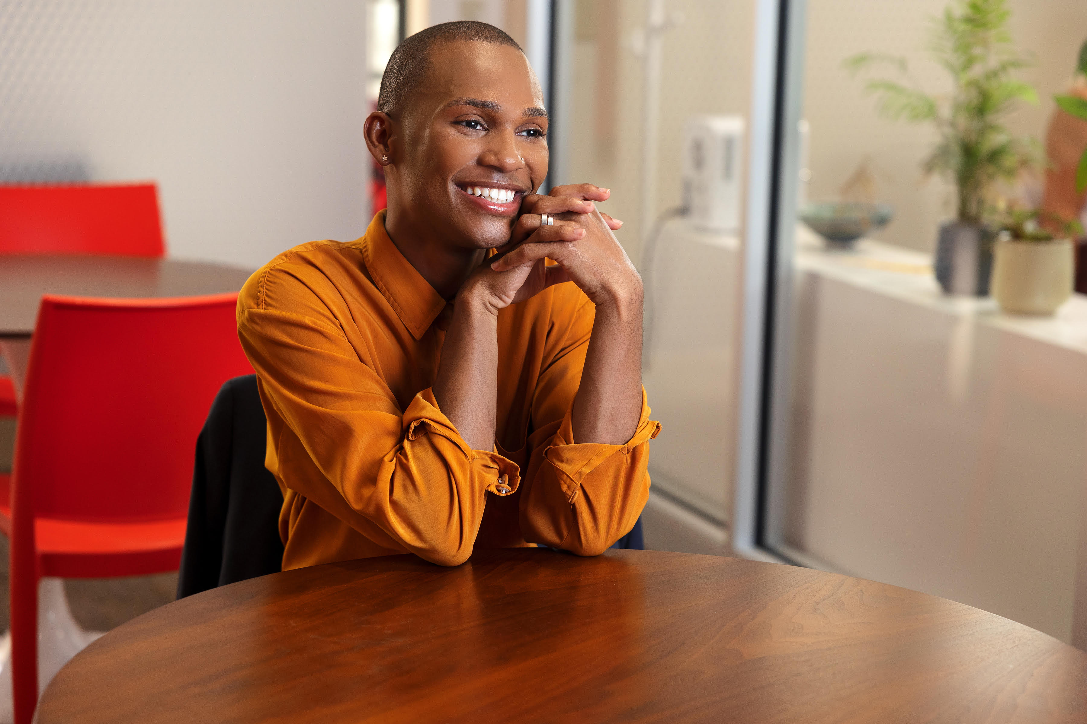 Dae Williams smiling, chin on folded hands, in a burnt-orange shirt at a wooden table